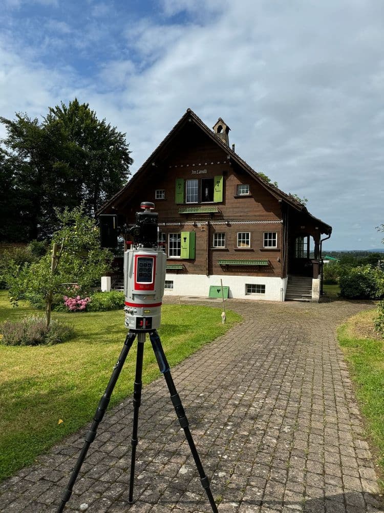 Vermessungsgerät auf Stativ vor traditionellem Holzhaus mit Garten.
